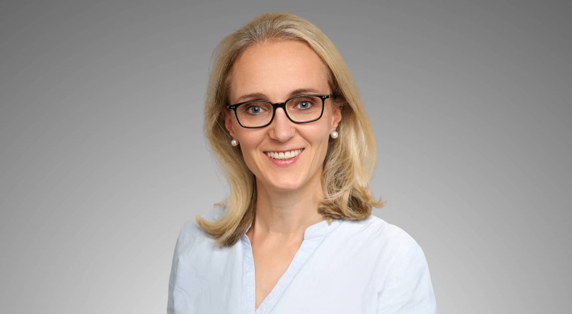 Eine Frau mit blondem Haar und Brille, die eine hellblaue Bluse trägt, lächelt in die Kamera vor einem schlichten grauen Hintergrund.